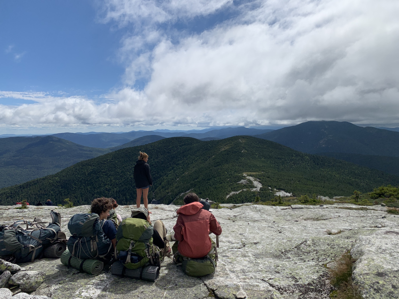 The image shows a group of people resting on a rocky mountaintop, enjoying a scenic view of rolling green hills and a partly cloudy sky. Some are sitting with their backpacks, while one person is standing and looking out at the landscape. The scene suggests a hiking or camping trip.